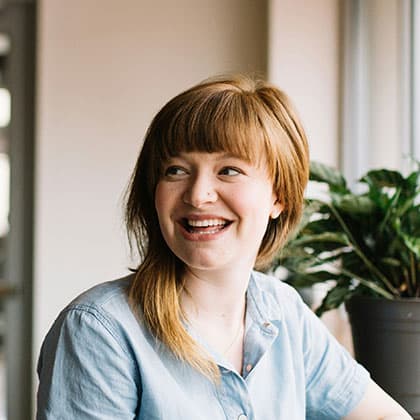 Happy person with short hair smiling, sitting indoors near a potted plant, wearing a light blue shirt, conveying a sense of satisfaction and positivity.